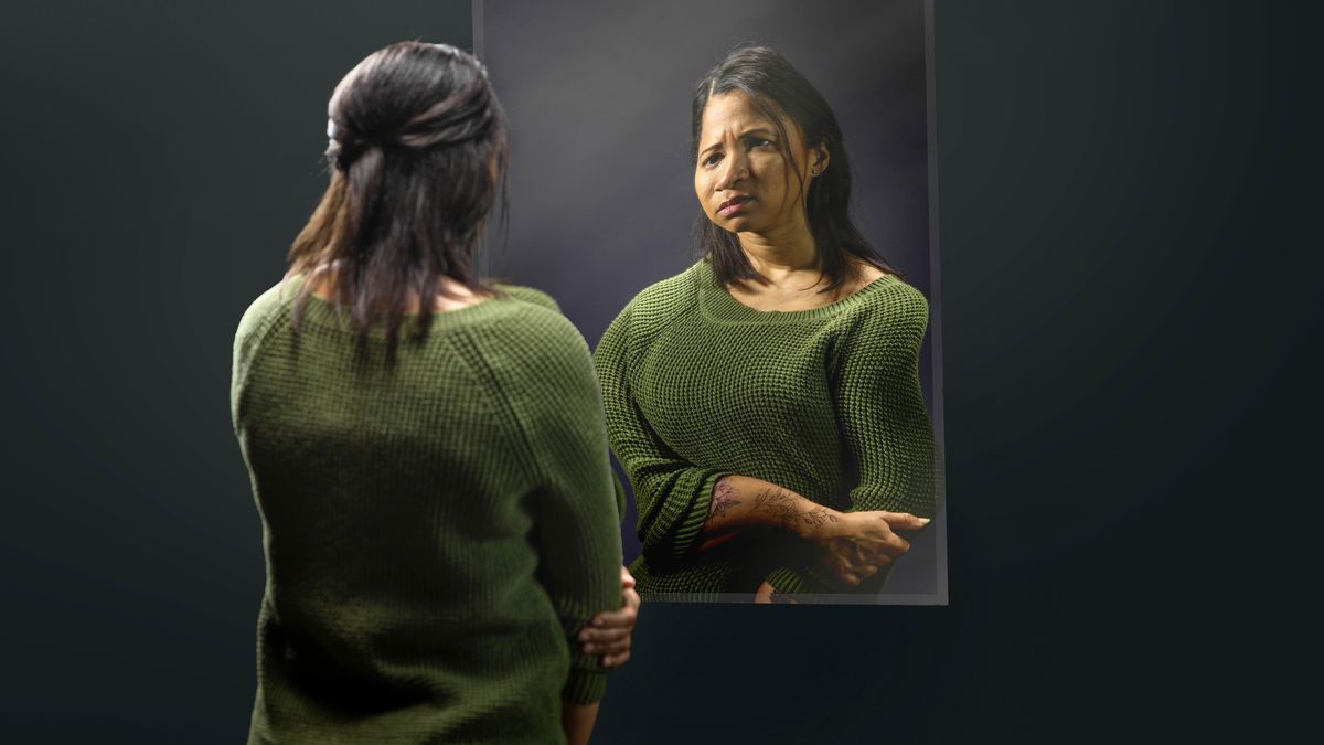 A discouraged sister with tattoos looks into a mirror that distorts her reflection.