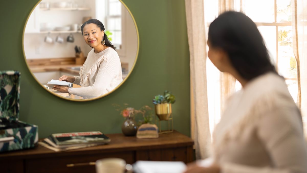 The sister from a previous image smiles while holding an open Bible and looking at her clear reflection in a mirror.